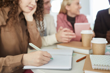 Close up of young woman writing in notebook while studying in college library with friends in background, copy space
