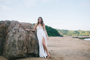Pretty content woman in white dress resting by rock at beach