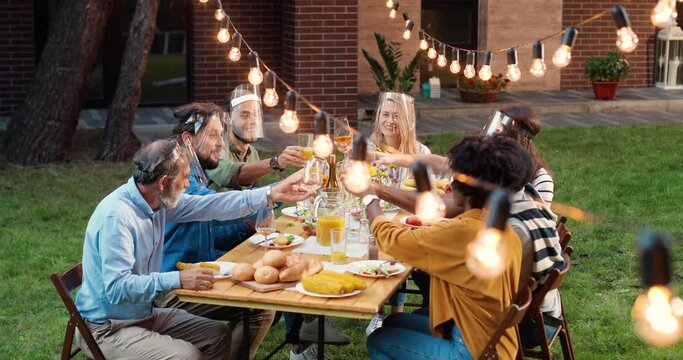 Mixed-races people in face shields sitting at dinner table in back yard, saying toasts and talking. Multi ethnic family having picnic meal in court and chatting friendly. Communication during pandemic