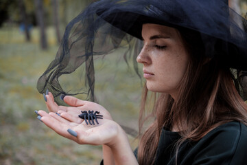 Young cute beautiful woman in dark dress and witch's hat holdsblack spider in her hands. Halloween party costume. Forest, park with autumn trees.