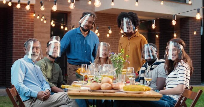 Portrait of mixed-races people in face shields at dinner table in back yard and smiling to camera. Multi ethnic family at picnic in court outdoors. Pandemic communication of generations.