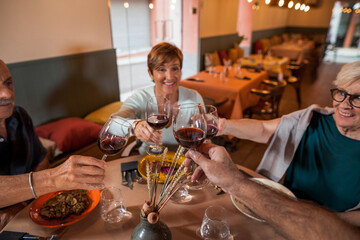 Senior friends group celebrating at dinner at restaurant. Detail of hands while toasting with glasses of red wine. Elderly lifestyle, friendship, food and drink, retired and pensioners concept