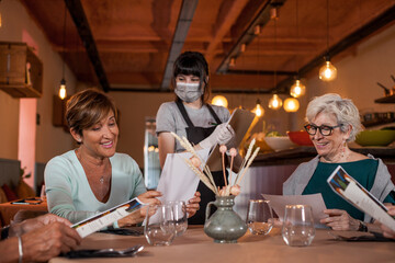 Two happy senior women with the menu in hands ordering food at a restaurant. Young waitress with protective face mask and gloves taking an order. Food and drinks during Coronavirus outbreak. 