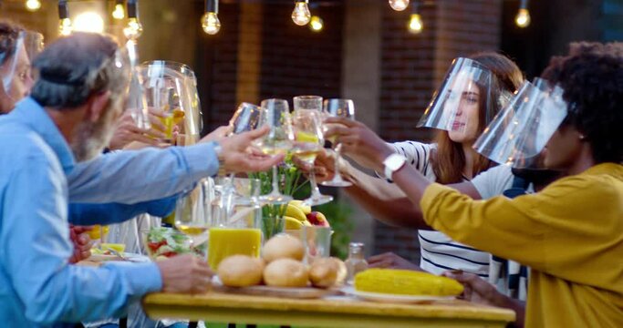 Happy multi ethnic family in medical face shields sitting at table with meal outdoor at picnic and toasting with juice and wine. Joyful young and old people having dinner and toast at barbrque.