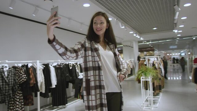 Pretty young woman is taking a selfie wearing a plaid coat at clothing store, tracking shot, 360 degree