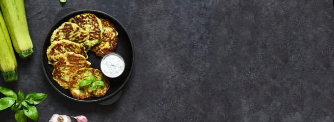 Zucchini pancakes with sauce and garlic in a frying pan on a black background. View from above.