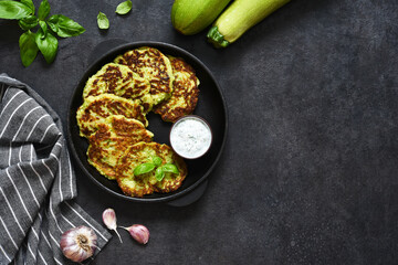 Zucchini pancakes with sauce and garlic in a frying pan on a black background. View from above.