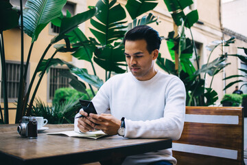 Focused ethnic man text messaging in cellphone while working in cafe terrace