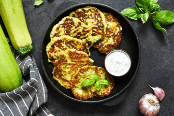 Zucchini pancakes with sauce and garlic in a frying pan on a black background. View from above.