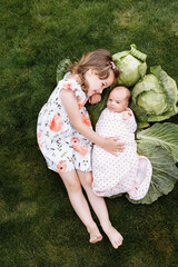 cute little girl with her younger newborn sister are lying on grass with cabbages outdoors. Relations between relatives. Two sisters love each other's. family lifestyle