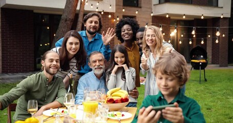 Mixed-races happy family at party dinner outdoor in yard smiling and posing to smartphone camera while small boy taking selfie photo. Multi ethnic people making photos together at barbeque Celebration - Powered by Adobe