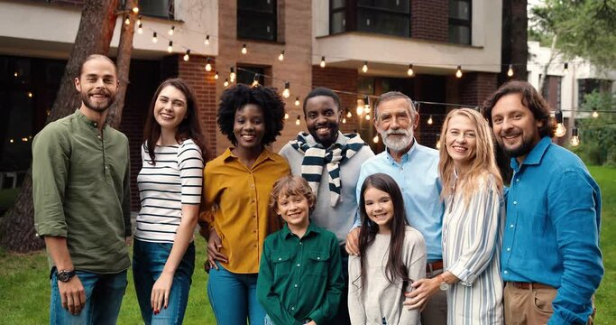 Portrait of mixed-races happy family at dinner outdoor in yard smiling and posing to camera on weekend spending together. Multi ethnic people at barbeque. Celebration. Parents and kids. Generations.