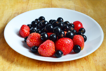 strawberries and black currants on a white plate and wooden table, harvesting, fresh and healthy vitamin-rich food