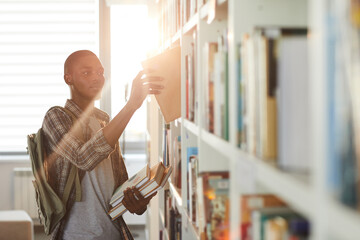 Waist up portrait of young African-American man taking book off shelf in school library against...