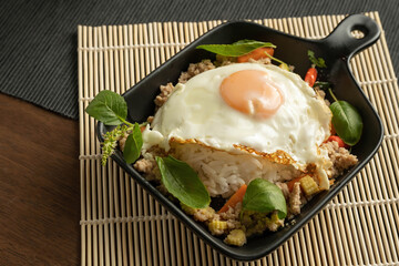 Stir-fried Thai basil with minced pork meat, carrots, and baby corns, with fried egg and fresh Thai basils sort in a black small pan on bamboo placemat, grey placemat, brown wood table. Copy space