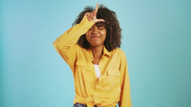 Young ethnic black lady laughing and showing loser hand gesture while posing against blue studio background