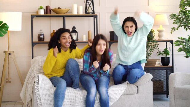 Female fans sitting on sofa in living room and watching sports match on TV. Excited women jumping and screaming when favorite team scoring goal. Concept of entertainment