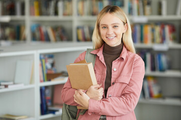 Waist up portrait of blonde young woman in school library holding books and smiling happily at...