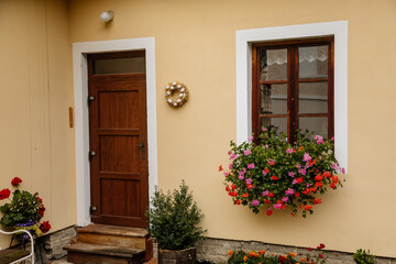 Fototapeta na wymiar Brown wooden door and vintage window with a red and pink flowers in a pot on a beige wall. Typical beautiful streets of the ancient city of Czech Republic. Entrance of an apartment or house