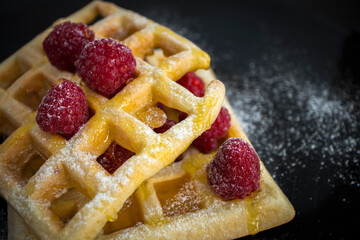 Two stacked waffles garnished with raspberries and me, sprinkled with powdered sugar on a black background. Food, breakfast. Closeup.
