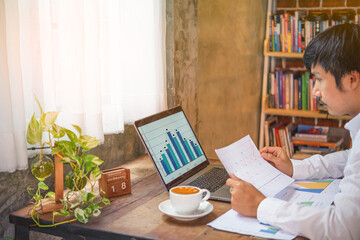 A man was working from home with a laptop ,paper sheet while  watching the graph statistic on the computer screeen.