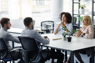 Team building and coffee break during work after quarantine. Multiracial workers with cups smiling and communicating through protective glass in interior