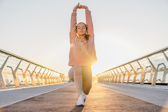 Young bruette woman with sport body warming up outdoors, stretching legs and hands preparing for running.