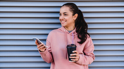 Close up photo of a young beautiful girl holding her phone and coffee in front of siding wall