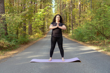 Fat caucasian woman with long curly dark hair dressed in black tshirt and black sportive pants is doing yoga on purple mat on the road in the autumn forest. Namaste.