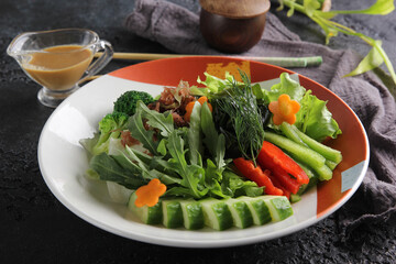 Japanese cuisine. Raw sliced vegetables: cucumber, pepper, carrot and broccoli. Arugula, lettuce, dill and sauce on a plate on a black table with chopsticks. Background image, copy space