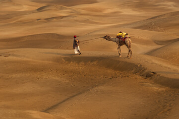 Man with camel walking across sand dunes in Jaisalmer, Rajasthan, India.