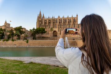 Young light-skinned girl taking a picture of the cathedral of Palma de Mallorca with her phone on her holidays. © MinekPSC