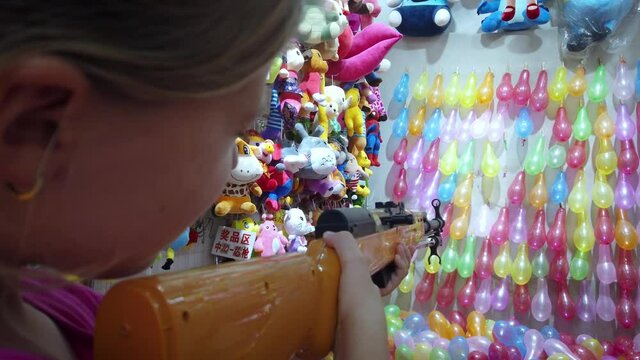 Young Caucasian girl shooting rubber bullet gun at rows of colorful balloons to win prizes at shooting range