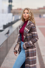 Elegant woman walking city street. Fashion fall autumn look. Pretty caucasian blonde stylish girl wearing blue jeans, wine red pullover and squared wool coat. Female with makeup and wavy blonde hair