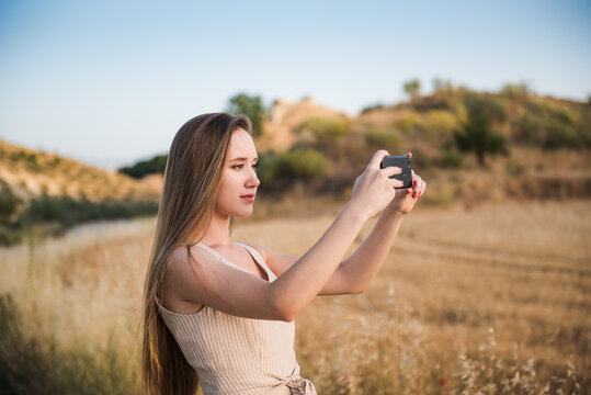 Young blonde woman taking photos in the field with her mobile phone. Caucasian girl traveling alone enjoys sunset in the field.