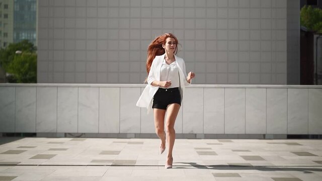 A beautiful woman with long red hair in a white coat runs in high heels along the city street. A pretty playful lady is running in casual clothes. Slow motion