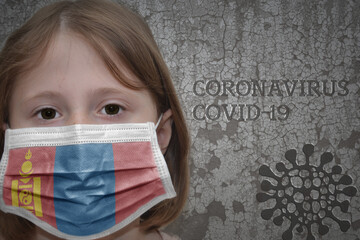 Little girl in medical mask with flag of mongolia stands near the old vintage wall with text coronavirus, covid, and virus picture. Stop virus concept