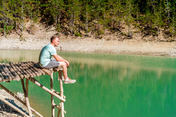 Fototapeta na wymiar Younf blond man on a bridge above a mountain lake with clear water and a view of a green forest. Copy space