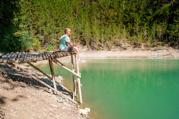 Younf blond man on a bridge above a mountain lake with clear water and a view of a green forest. Copy space