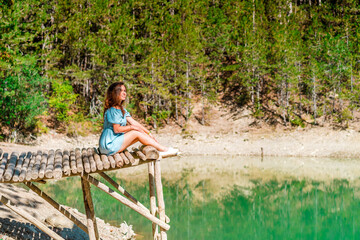 Fototapeta na wymiar Beautiful girl with long hair in a dress on a bridge above a mountain lake with clear water and a view of a green forest. Copy space