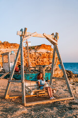 Romantic evening at sunset by the sea, a young woman in a dress with a bouquet of flowers is sitting on a wooden swing holding her hat