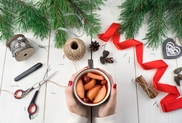 Break with a Cup of mulled wine while preparing for the Christmas holidays. Women's hands hold a mug with a drink on the background of pine branches, red ribbon and scissors. New year and Christmas.