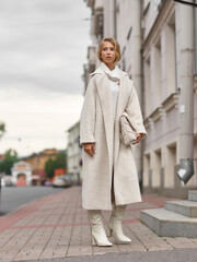 Fototapeta premium Elegant woman in white dress, hessian boots and coat walking at city street. Fall autumn fashion look. Pretty tall stylish young gitl with fashionable makeup and hair style. Elegant lady. Full length