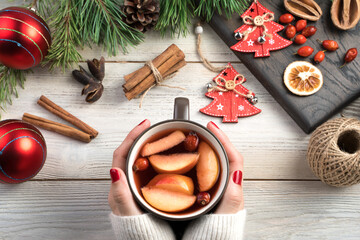 Women's hands hold a mug of mulled wine on a white wooden background surrounded by Christmas toys, berries, cinnamon and pine branches. The concept of New year and Christmas.