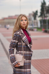 Fototapeta premium Elegant woman walking city street. Fashion fall autumn look. Pretty caucasian blonde stylish girl wearing blue jeans, wine red pullover and squared wool coat. Female with makeup and wavy blonde hair