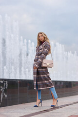 Elegant woman walking city street. Fashion fall autumn look. Pretty caucasian blonde stylish girl wearing blue jeans, wine red pullover and squared wool coat. Female with makeup and wavy blonde hair