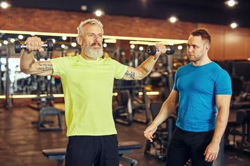 Fotobehang Persoonlijk Training body in any age. Focused mature caucasian man in sportswear exercising with dumbbells under supervision of a young professional personal trainer at gym  © Kostiantyn