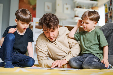 Father with sons reading book, spending time together at home and lying on the floor