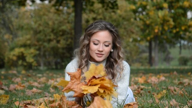 A young caucasian girl in an autumn park lies on the leaves and smiles with a leaf in her hand