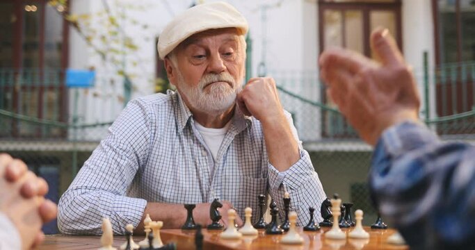 Portrait shot of the Caucasian grey haired good looking man on retirement sitting at the table on a fresh air, leaning on his arm and looking at the friend's hand moving a chessman on a board